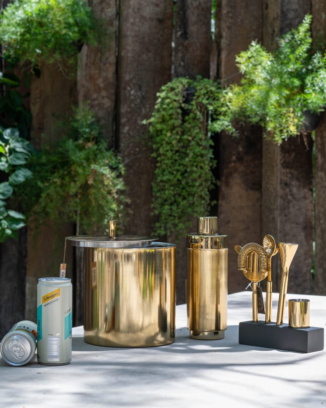Kit bar dourado da Maria Pia Casa — balde de gelo, coqueteleira e utensílios — fotografado ao ar livre.